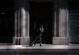 A man in a suit walks along a sunlit pavement, pulling a suitcase behind him. The scene is set against the backdrop of a large, dark wooden door flanked by stone walls. Partial shadows create a dramatic contrast, highlighting the architectural details. Signs are visible on the right side, written in different languages.