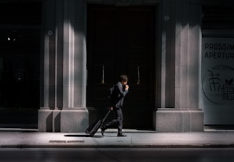A man in a suit walks along a sunlit pavement, pulling a suitcase behind him. The scene is set against the backdrop of a large, dark wooden door flanked by stone walls. Partial shadows create a dramatic contrast, highlighting the architectural details. Signs are visible on the right side, written in different languages.