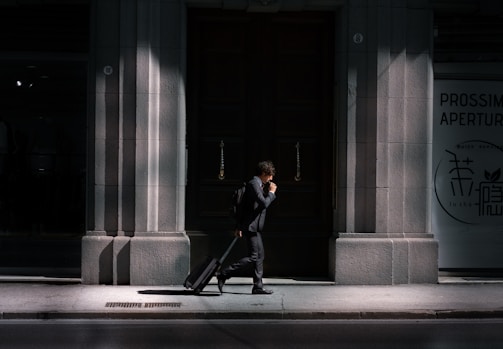 A man in a suit walks along a sunlit pavement, pulling a suitcase behind him. The scene is set against the backdrop of a large, dark wooden door flanked by stone walls. Partial shadows create a dramatic contrast, highlighting the architectural details. Signs are visible on the right side, written in different languages.