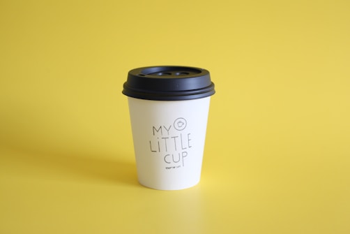 A paper coffee cup with a black lid is placed against a bright yellow background. The cup has the text 'My Little Cup' printed on it.