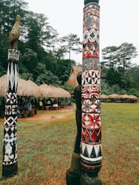 Colorful carved wooden poles stand prominently in a grassy area, featuring intricate patterns with red, white, and black designs. Behind the poles, several traditional thatched huts are partially visible, surrounded by dense, lush green trees.