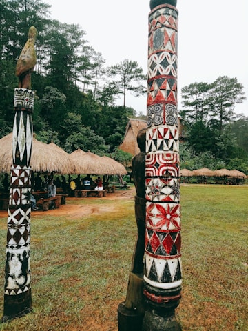 Colorful carved wooden poles stand prominently in a grassy area, featuring intricate patterns with red, white, and black designs. Behind the poles, several traditional thatched huts are partially visible, surrounded by dense, lush green trees.