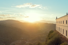 A golden sunset over rolling hills, casting warm light across a lush green landscape. A large, elegant building with multiple windows stands at the edge of a hill on the right side, adding a sense of scale and architecture to the serene natural view.