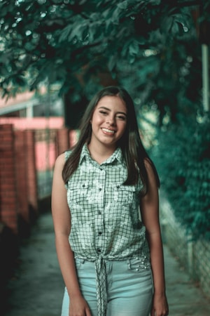 A young woman stands outdoors, smiling warmly. She is wearing a sleeveless plaid shirt and light-colored jeans. The background features greenery with trees and a brick wall, creating a natural and serene environment.