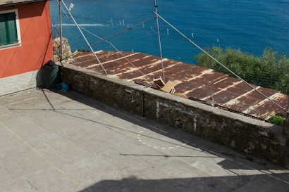 A rooftop patio with a rusted metal roof adjacent to a vibrant orange building. The patio is enclosed by a stone wall. In the background, there is a view of the ocean with clear blue water and greenery lining the horizon. Some boxes and containers are placed near the wall.