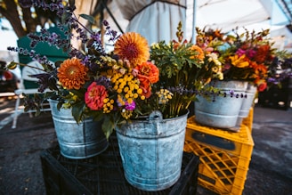 Metal buckets filled with vibrant, colorful flowers are arranged on wooden crates at an outdoor market setting. The floral arrangements include a variety of flowers such as orange and red dahlias, yellow billy buttons, purple lavender, and other mixed greens. The setting is under a large canopy, with a parking lot visible in the background.