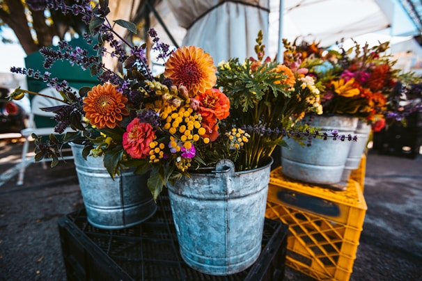 Metal buckets filled with vibrant, colorful flowers are arranged on wooden crates at an outdoor market setting. The floral arrangements include a variety of flowers such as orange and red dahlias, yellow billy buttons, purple lavender, and other mixed greens. The setting is under a large canopy, with a parking lot visible in the background.