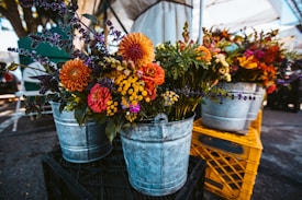 Metal buckets filled with vibrant, colorful flowers are arranged on wooden crates at an outdoor market setting. The floral arrangements include a variety of flowers such as orange and red dahlias, yellow billy buttons, purple lavender, and other mixed greens. The setting is under a large canopy, with a parking lot visible in the background.