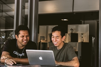Two people are sitting at a table indoors, both smiling and looking at a laptop screen. They appear to be engaged and enjoying what they are viewing. Behind them, there is a shelf with decorative items and neutral lighting.
