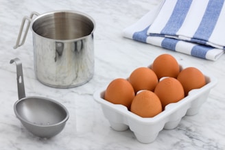 A stainless steel pot is placed on a marble countertop next to a set of brown eggs in a white ceramic egg holder. A metal ladle lies in front of the pot. The background features a folded blue and white striped cloth.