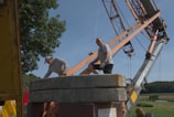 Two people are working around a large industrial crane set up in a rural area. One person is wearing a cap and leaning over, while the other wears sunglasses and stands next to heavy cement counterweights. The crane's metal arm and the lush green landscape in the background are visible.