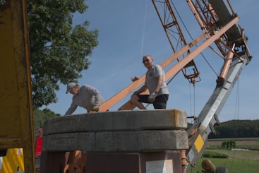 Two people are working around a large industrial crane set up in a rural area. One person is wearing a cap and leaning over, while the other wears sunglasses and stands next to heavy cement counterweights. The crane's metal arm and the lush green landscape in the background are visible.