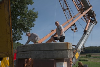 Two people are working around a large industrial crane set up in a rural area. One person is wearing a cap and leaning over, while the other wears sunglasses and stands next to heavy cement counterweights. The crane's metal arm and the lush green landscape in the background are visible.
