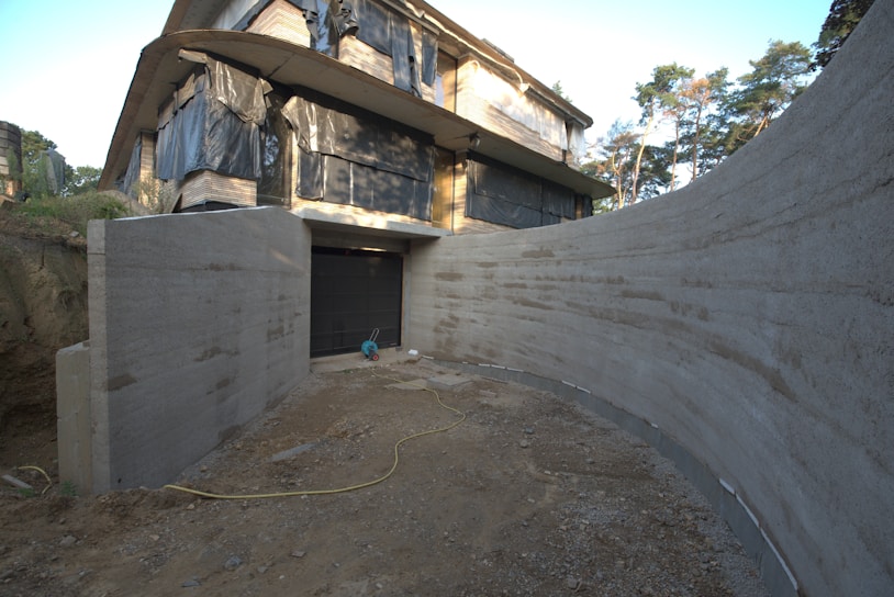 A partially constructed building with large sections of exposed concrete and black coverings over the windows sits amid trees. The ground in front appears unfinished with a dirt path leading to a closed garage door. A yellow electrical cord is visible on the ground, and construction materials are scattered around.