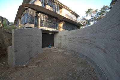 A partially constructed building with large sections of exposed concrete and black coverings over the windows sits amid trees. The ground in front appears unfinished with a dirt path leading to a closed garage door. A yellow electrical cord is visible on the ground, and construction materials are scattered around.
