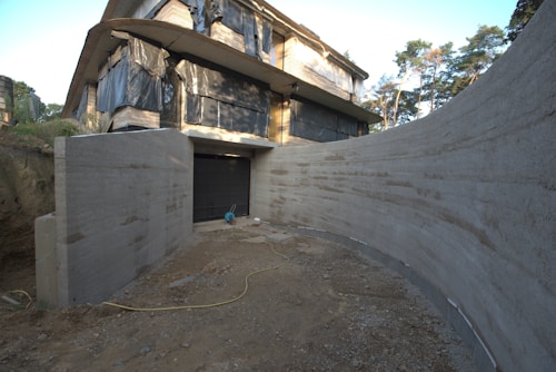 A partially constructed building with large sections of exposed concrete and black coverings over the windows sits amid trees. The ground in front appears unfinished with a dirt path leading to a closed garage door. A yellow electrical cord is visible on the ground, and construction materials are scattered around.