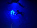 Three divers are submerged underwater in a pool. The image is illuminated with a striking blue light, giving an ethereal glow to the water. Two divers are near the surface holding onto equipment or a platform, while the third diver is slightly deeper, possibly adjusting something. A rope is also visible extending into the water.
