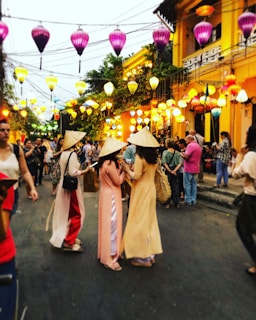 A lively street scene with colorful lanterns hanging above, illuminating the surroundings. People are gathered, some in traditional attire with conical hats, adding a cultural and festive feel. The street is bustling with activity, as groups of people interact and enjoy the vibrant atmosphere.