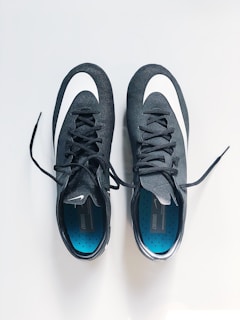 A pair of black sports shoes with white swoosh logos are placed side by side on a clean, light-colored surface. The insoles are blue with a pattern of small black dots, and the laces are neatly tied.