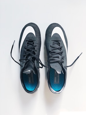 A pair of black sports shoes with white swoosh logos are placed side by side on a clean, light-colored surface. The insoles are blue with a pattern of small black dots, and the laces are neatly tied.