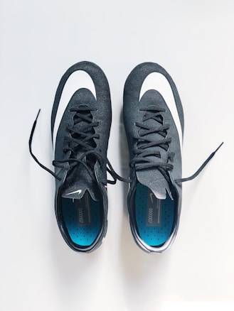 A pair of black sports shoes with white swoosh logos are placed side by side on a clean, light-colored surface. The insoles are blue with a pattern of small black dots, and the laces are neatly tied.