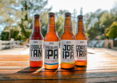 Four beer bottles are lined up on a wooden table outdoors. The labels on each bottle read Goggle Tan, Apocalypse IPA, Joe IPA, and Coast Trip. The sun casts a warm glow on the bottles, and a blurred natural background suggests a sunny day.