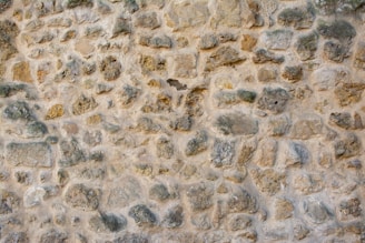 A textured pattern of natural stone and mortar arranged to form a wall. The stones are irregular in shape and size, with varying shades of beige, gray, and brown.