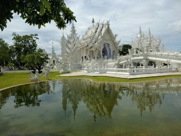 An intricately designed white temple with elaborate carvings stands adjacent to a reflective pond. The temple's details are mirrored in the water, surrounded by lush green grass and trees. The sky is partially cloudy, adding to the serene ambiance.