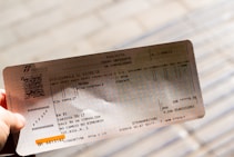 A hand holding an Italian train ticket from Trenitalia. The ticket features detailed text including departure and arrival locations, dates, and a barcode. The background is blurred, focusing attention on the ticket.