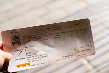 A hand holding an Italian train ticket from Trenitalia. The ticket features detailed text including departure and arrival locations, dates, and a barcode. The background is blurred, focusing attention on the ticket.