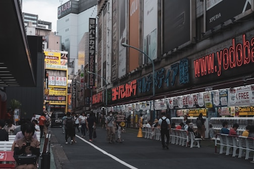 A bustling urban street with numerous people walking and sitting. The area is lined with buildings covered in colorful advertisements and signage, featuring both digital and printed displays. Neon lights and billboards add a vibrant atmosphere to the scene.