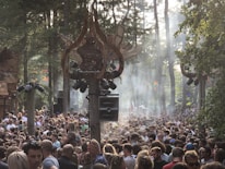 A large crowd of people gathered in a forest setting with trees surrounding them. Sunlight is streaming through the trees, creating a hazy and atmospheric effect. Architectural structures with artistic designs and large speakers are visible above the crowd, suggesting a music festival or outdoor event.