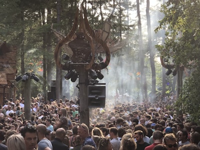 A large crowd of people gathered in a forest setting with trees surrounding them. Sunlight is streaming through the trees, creating a hazy and atmospheric effect. Architectural structures with artistic designs and large speakers are visible above the crowd, suggesting a music festival or outdoor event.