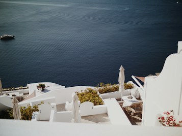 White architecture of a coastal setting with terraces overlooking a deep blue sea. The scene includes sun loungers, white parasols, and vibrant greenery. A distant yacht is seen on the calm water, adding a sense of tranquility.