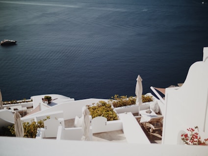 White architecture of a coastal setting with terraces overlooking a deep blue sea. The scene includes sun loungers, white parasols, and vibrant greenery. A distant yacht is seen on the calm water, adding a sense of tranquility.