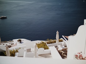 White architecture of a coastal setting with terraces overlooking a deep blue sea. The scene includes sun loungers, white parasols, and vibrant greenery. A distant yacht is seen on the calm water, adding a sense of tranquility.