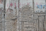 A section of a concrete wall featuring worn surfaces and several vertical metal rods embedded in the structure. The wall has visible signs of erosion and weathering, with various graffiti and faded paint in red and blue colors. The concrete shows patches of rough texture and exposed aggregate.