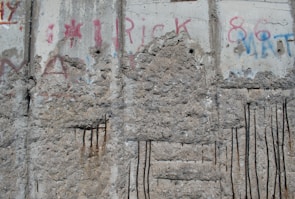 A section of a concrete wall featuring worn surfaces and several vertical metal rods embedded in the structure. The wall has visible signs of erosion and weathering, with various graffiti and faded paint in red and blue colors. The concrete shows patches of rough texture and exposed aggregate.
