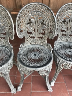 Three intricately designed metal chairs with ornate floral patterns and circular decorative elements. The chairs have a vintage, artistic style and are positioned on a red tiled floor against a wooden backdrop.