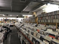 A spacious, well-lit record store with rows of vinyl records organized on long tables and shelves. The walls are lined with additional stacks of records. Several categories like rock and folk are distinguished by labels on dividers. A person is seen in the distance browsing the collection.