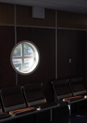 A dimly lit waiting area with a row of empty chairs. A circular porthole window provides a view of some trees outside, allowing natural light to partially illuminate the seats.