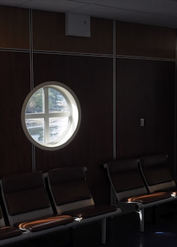 A dimly lit waiting area with a row of empty chairs. A circular porthole window provides a view of some trees outside, allowing natural light to partially illuminate the seats.