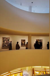 A modern art museum interior features a spiral ramp with people observing colorful abstract paintings on white walls. The architecture includes smooth curved lines and warm lighting, creating an inviting atmosphere.