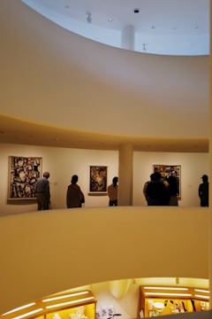 A modern art museum interior features a spiral ramp with people observing colorful abstract paintings on white walls. The architecture includes smooth curved lines and warm lighting, creating an inviting atmosphere.