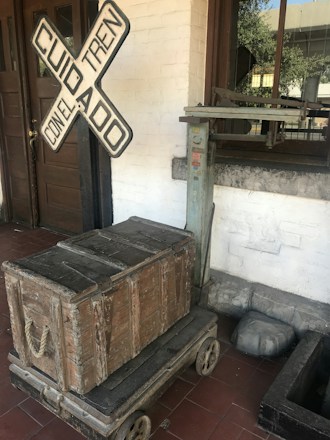 A vintage wooden crate sits on a wheeled platform in front of a building with a wooden door. A railway crossing sign with the words 'Cuidado Con El Tren' is leaning against the wall. There is also an old weighing scale near the window, adding to the historical or rustic ambiance.