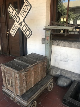 A vintage wooden crate sits on a wheeled platform in front of a building with a wooden door. A railway crossing sign with the words 'Cuidado Con El Tren' is leaning against the wall. There is also an old weighing scale near the window, adding to the historical or rustic ambiance.