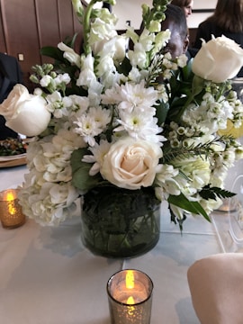 A floral arrangement featuring white roses, daisies, and greenery placed in a transparent vase. The setting appears to be a formal dining table with candles, some lit, and a white tablecloth. Several people are partially visible in the background.