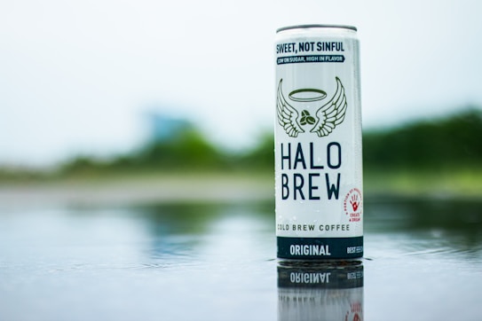 A can of Halo Brew cold brew coffee stands upright on a reflective surface. The can features a design with wings and a halo, underlined by the slogan 'Sweet, Not Sinful'. A natural and blurred background presents a soft and serene atmosphere.