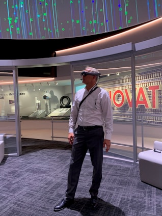 A man in formal attire stands in a modern, technology-themed space. He is wearing augmented reality glasses and is surrounded by displays and panels showcasing innovative concepts. The ceiling displays an array of digital patterns with numeric and symbolic designs.