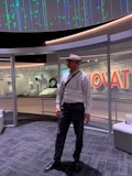 A man in formal attire stands in a modern, technology-themed space. He is wearing augmented reality glasses and is surrounded by displays and panels showcasing innovative concepts. The ceiling displays an array of digital patterns with numeric and symbolic designs.